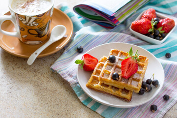 Background breakfast. A cup of cappuccino coffee and Belgian waffles with berries on the kitchen table.