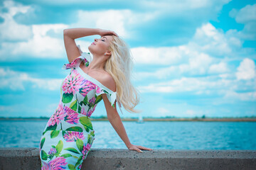 Obraz na płótnie Canvas Beautiful blonde hair woman in romantic flowers dress near blue water and sand. Cute happy girl have fun on a nature