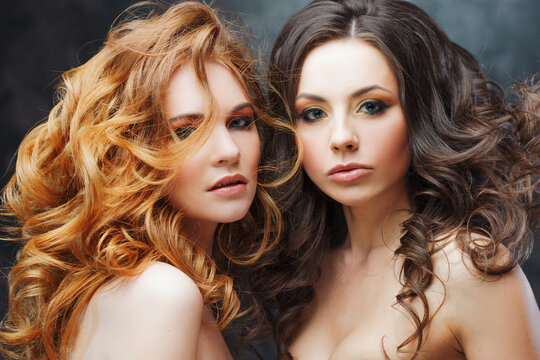 Two charming beauties with luxurious curly hair. Brunette and redhead. Studio portrait on the dark background.