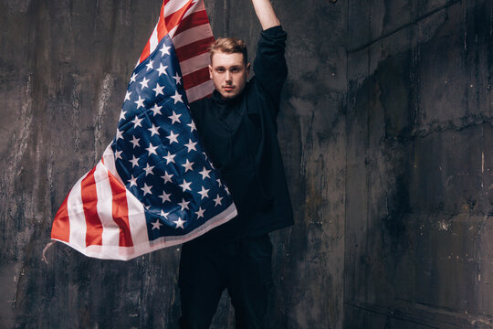 Young USA Patriot With Flying National Flag. Strong Man In Black Cloth On Dark Studio Background. Independence Day, Confidence, Pride, Fidelity To The Nation, Memorial Day Concept