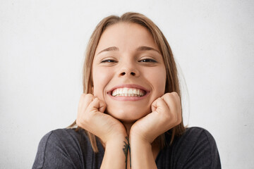 Close up indoor portrait of happy excited young female celebrating success and promotion at work, looking at camera with broad cheerful smile, holding hands on her face, feeling joy and happiness