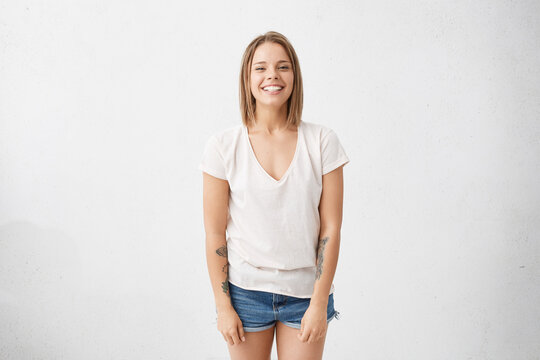 Youth and happiness. Style and fashion. Isolated studio shot of attractive fashioable cheerful young female wearing white t-shirt and jeans shorts smiling broadly, happy with good positive news