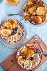 Homemade pancakes with banana and black currant on painted ceramic plates, with wooden board and tablecloth, with vintage fork and spoon. Flatlay food photo.