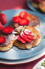 Ukrainian Syrniki or homemade cheesy pancakes with mint and strawberry on beige and blue ceramic plates on red tablecloth with forks.