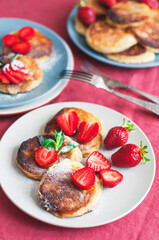 Ukrainian Syrniki or homemade cheesy pancakes with mint and strawberry on beige and blue ceramic plates on red tablecloth with forks.