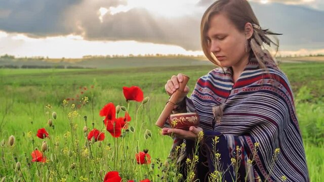 Young beautiful woman in poncho with ethnic pattern sits on field in the middle of green grass and red poppies at the background of cloudy sunset sky and plays tibetan singing bowl.
