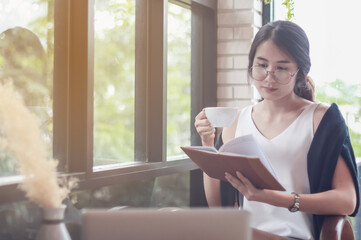 woman reading a book at the wooden counter desk in coffee shop with smile face in happy motion on blurred coffee shop view background under window light