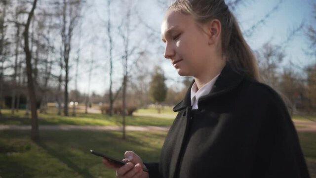 Gimbal shot of female teen girl using smartphone and walking in town park in spring sunny day, uhd prores footage