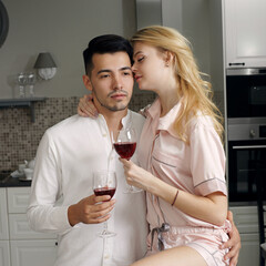 Young caucasian couple sitting at home ktichen, hugging, smiling, drinking red wine. Young lovers having good time. Blonde woman and brunette man