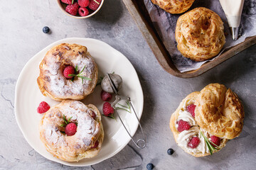 Filling and empty homemade choux pastry cake Paris Brest with raspberries, almond, sugar powder, rosemary on plate and oven tray with berries over gray texture background. French dessert. Flat lay