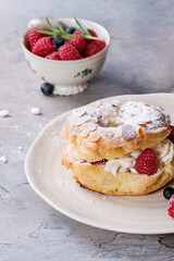 Homemade choux pastry cake Paris Brest with raspberries, almond, sugar powder, rosemary on white plate with berries over gray texture background. French dessert.