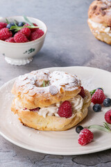 Homemade choux pastry cake Paris Brest with raspberries, almond, sugar powder, rosemary on white plate with berries over gray texture background. French dessert.