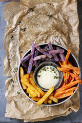 Variety of french fries traditional potatoes, purple potato, carrot served with white cheese sauce, salt, thyme in bowl on baking paper gray metal background. Top view with space. Homemade fast food