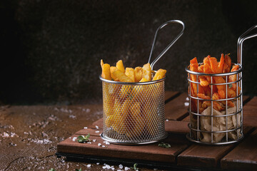 Variety of french fries traditional potatoes, sweet potato, carrot served in frying basket with salt, thyme on wooden board over brown texture background. Homemade fast food