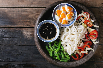 Ingredients for cooking stir fry udon noodles, green beans, sliced paprika, boiled eggs, soy sauce with sesame seeds in traditional bowls in terracotta tray over old wooden background. Flat lay
