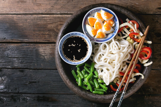 Ingredients for cooking stir fry udon noodles, green beans, sliced paprika, boiled eggs, soy sauce with sesame seeds in traditional bowls with wooden chopsticks over old wooden background. Flat lay