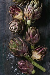 Uncooked whole and sliced organic wet purple artichokes with salt over dark wooden background. Rustic style. Top view with space