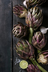 Uncooked whole and sliced organic wet purple artichokes with salt, lime and garlic over dark wooden background. Rustic style. Top view with space