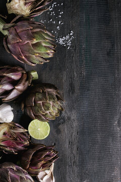 Uncooked whole and sliced organic wet purple artichokes with salt, lime and garlic over dark wooden background. Rustic style. Top view with space
