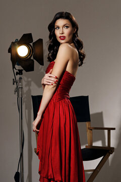 Young pretty beautiful woman in red long evening dress with makeup, red lips and classical hollywood waves hairstylestanding at film set with two cinema lights.