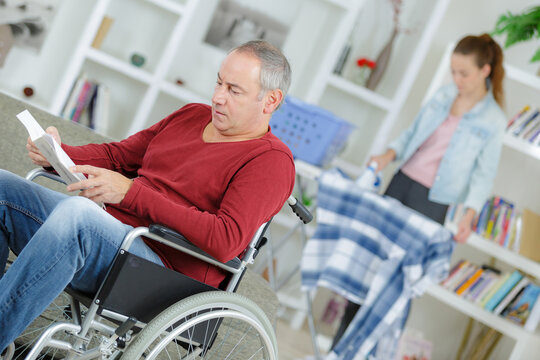 Man in wheelchair reading book, carer ironing in background