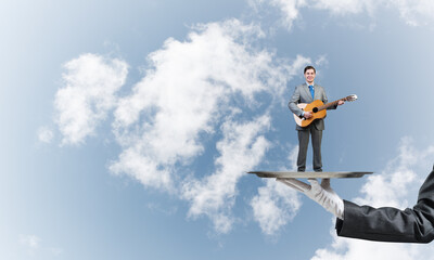 Businessman on metal tray playing acoustic guitar against blue sky background