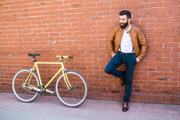 Fototapeta na wymiar Young stylish man with beard in a brick background standing near the bike with bag in hands