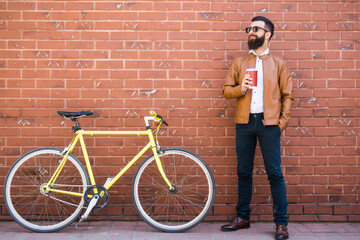 Young bearded man drinking coffee while sitting near his bicycle outdoors