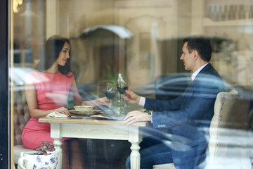 Beautiful romantic couple having dinner with wine in bright luxury restaurant. View through window from outside
