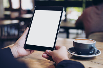 Mock up image of business woman's hands holding black tablet with white blank screen and coffee cup on wooden table in cafe
