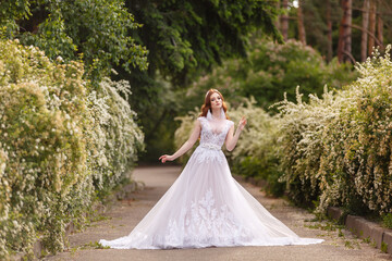 Beautiful redhead Bride in fantastic wedding dress in blooming garden. Growth Portrait in sunset light. Pretty young caucasian redhead girl walks in a garden and plays with her dress. Young princess