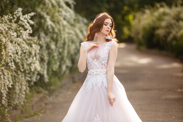 Fototapeta na wymiar Beautiful redhead Bride in fantastic wedding dress in blooming garden. Portrait in sunset light. Pretty young caucasian redhead girl walks in a garden and plays with her dress. Young princess