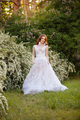 Obraz na płótnie Canvas Beautiful redhead Bride in fantastic wedding dress in blooming garden. Growth Portrait in sunset light. Pretty young caucasian redhead girl walks in a garden and plays with her dress. Young princess