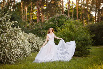 Beautiful redhead Bride in fantastic wedding dress in blooming garden. Growth Portrait in sunset light. Pretty young caucasian redhead girl walks in a garden and plays with her dress. Young princess