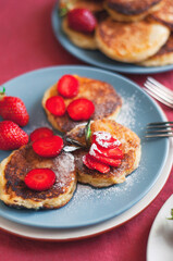 Ukrainian Syrniki or homemade cheesy pancakes with mint and strawberry on beige and blue ceramic plates on red tablecloth with forks.