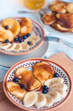 Homemade pancakes with honey, banana and black currant on painted ceramic plates, with wooden board and tablecloth, with vintage fork and spoon.