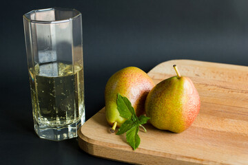 Soft non-alcoholic pear cider  in a transparent glass and two pears with fresh basil leaves on wooden cutting board with black background