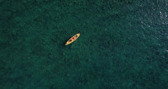 Silhouette of tiny blonde female in bright red one piece swimsuit resting on top of yellow kayak, cruisng through crystal clear waters of mediterranean sea