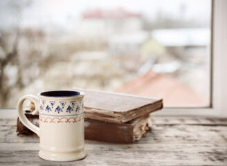 Patterned ceramic cup of hot tasty drink and old books on wooden windowsill