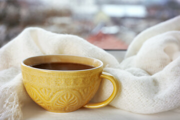 Patterned cup of hot tasty drink and fluffy scarf on windowsill