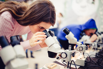 The student girl looks through a microscope in an equipped classroom with a series of microscopes. The concept is to study the science of biology, organisms, bacteria, viruses.