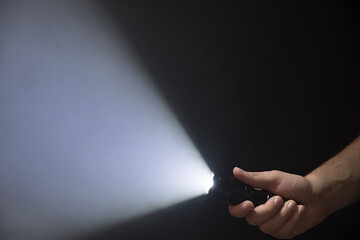 Black flashlight with a beam of light in male's hand isolated from right side of the frame on black background - obrazy, fototapety, plakaty