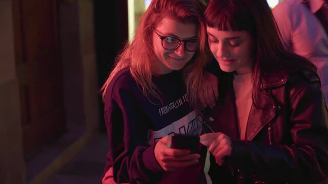Two best friends girls on night street with red light read comments to their story or photo post in social media they just made. They discuss replies and counts likes.