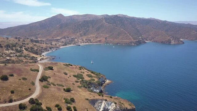 Aerial shot flying over hills along a rocky coastline on the Pacific ocean in California. Located at Two Harbors, California. Clear blue skies and water, several boats visible. Birds flying below.