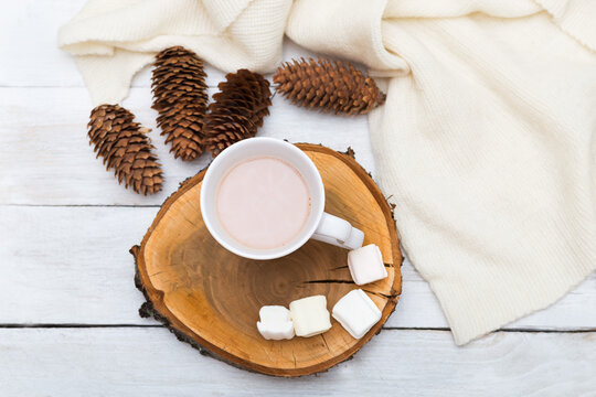 Cup with hot chocolate and marshmallows, a row of cones and a knitted blanket on a white wooden background