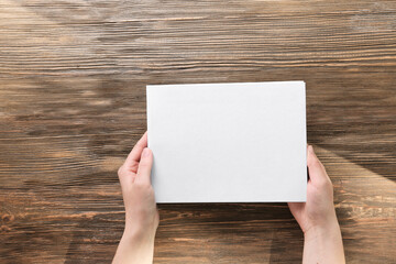 Obraz premium Female hands holding closed book with blank cover on wooden background