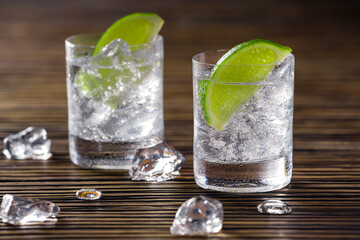Two glasses with gin and tonic served with ice and lime on a wooden table.