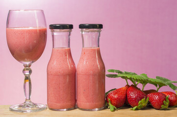 A glass and two bottles with strawberry smoothie on a pink background