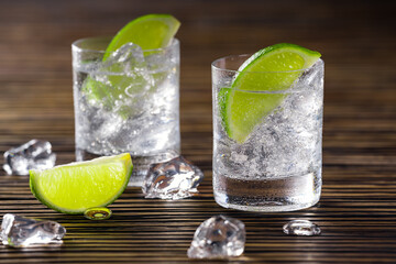 Two glasses with gin and tonic served with ice and lime on a wooden table.
