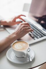 Cup of hot coffee or cappuccino with a pattern. Blurred hands are typing on keyboard of a laptop in the background.